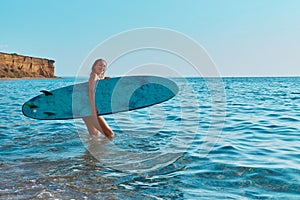 Happy girl walk in ocean with surfboard. Calm sea view on summer day. Idea of surf sport, beach day and summer holidays