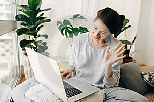 Happy girl waving hi to laptop, video chatting with family or friends, sitting in modern room with pillows and plants. Young