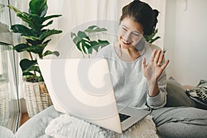 Happy girl waving hi to laptop, video chatting with family or friends, sitting in modern room with pillows and plants. Young
