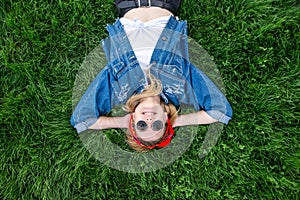 Happy girl wearing sunglasses and a jeans jacket lays on the grass, looks up and smiles