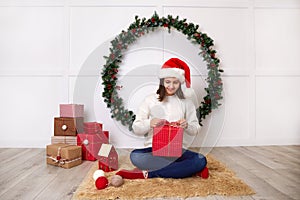 A happy girl in a white knitted sweater and a Santa Claus hat holds a gift box in her hands.