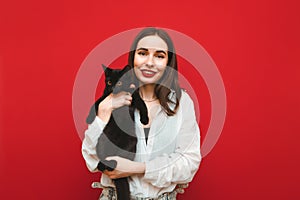 Happy girl in a white shirt stands on a red background with a black cat in her hands, looks into the camera and smiles. Smiling