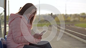 Happy girl writes message using her smart phone in the tram stop.