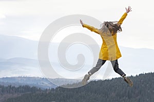 Happy girl in yellow jacket jumps with hands up to the sky on the top of mountains. Travel hiking and tourism