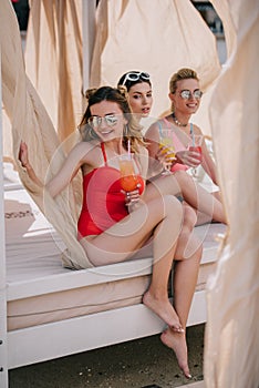 happy girlfriends in swimwear and sunglasses drinking cocktails in bungalow