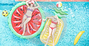 Happy girls floating with tropical fruit lilos inside swimming pool - Young women friends relaxing in summer vacation at resort
