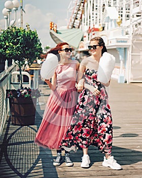 Happy girls with sunglasses and prom dresses holding cotton candy at amusement park. Teenagers in pink evening dresses on vacation