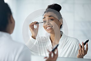 Happy glamour millennial girl looking in mirror, applying mascara