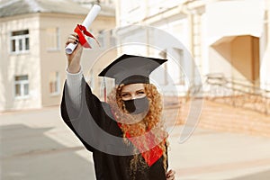 Happy grad student, she is in a black mortarboard with red tassel, and a face mask, in gown shows a diploma in her hand