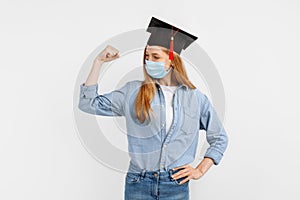 Happy graduate girl, wearing a medical protective mask on her face and a graduation cap on her head, raises her hand and shows her