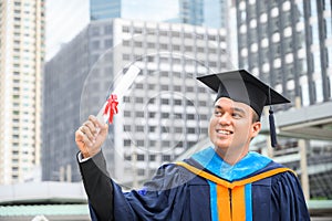 Happy graduate. Happy Asain man in graduation gowns holding diploma in hand on urban city background