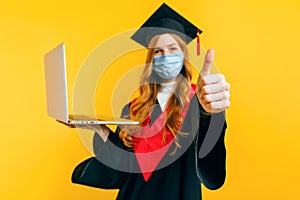 A happy, graduate, with a medical protective mask on her face, uses a laptop and gives a thumbs-up on a yellow background.