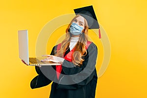 A happy, graduate, with a medical protective mask on her face, uses a laptop on an isolated yellow background. Distance learning,