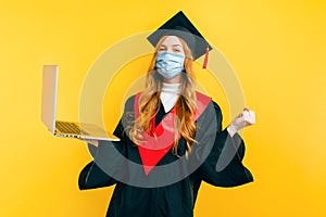 A happy, graduate, with a medical protective mask on her face, uses a laptop on an isolated yellow background. Distance learning,