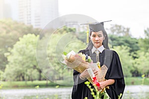 Happy graduate young Asian woman in cap and gown holding bouquet and certificated in hand, Education concept