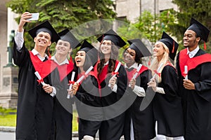 Happy graduates taking selfie together at university campus, using smartphone