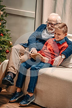 happy grandfather and grandson reading book on christmas while sitting