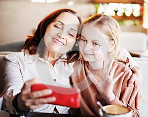 Happy grandmother and cute granddaughter taking selfies on the phone together, while sitting at the cafe