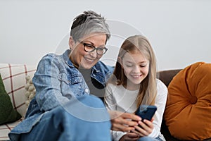 Happy grandmother and granddaughter watching fun content on phone in home interior