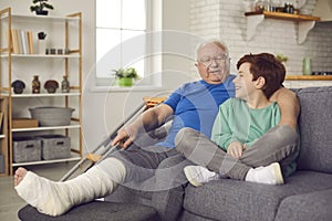 Happy grandson and his grandfather with broken leg in cast sitting on couch and talking