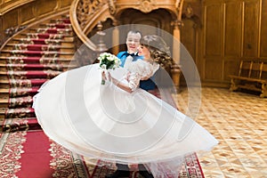 Happy groom carrying his wife with bouquet near big wooden stairs at old vintage house