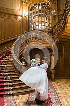 Happy groom carrying his wife with bouquet near big wooden stairs at old vintage house