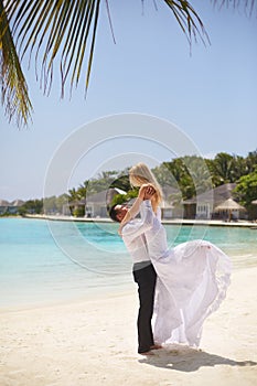 Happy groom holds bride on his hands under wedding ceremony arch on tropical island beach on Maldives. Turquoise ocean