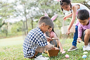 Happy group of diverse cute children hunting Easter eggs, girl wearing bunny ears. kids holding basket, picking eggs on grass