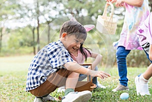 Happy group of diverse cute children hunting Easter eggs, girl wearing bunny ears. kids holding basket, picking eggs on grass