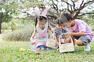 Happy group of diverse cute children hunting Easter eggs, girl wearing bunny ears. kids holding basket, picking eggs on grass