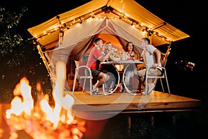 Happy group of friends relaxing in glamping and drinking wine on summer evening near cozy bonfire. Luxury camping tent for outdoor