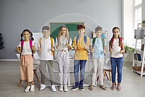 Happy group of schoolchildren ready for school showing thumbs up gesture in classroom.