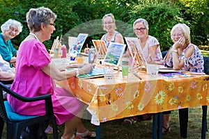 Happy group of senior ladies enjoying art class seated around a table outdoors in the garden painting with water colors while