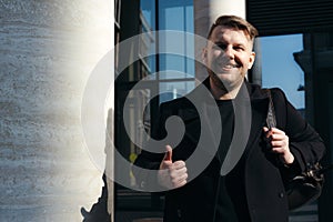 Happy guy with backpack on shoulder showing thumb up smiling at camera while standing on mirrored building photo