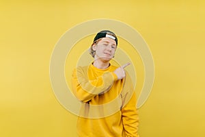 Happy guy in a cap and sweatshirt stands on a yellow background and points his finger to the side with a smile on his face