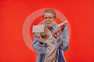 Happy guy with closed eyes with a beard in a blue shirt rejoices at the gift that is in an open white box with a red bow