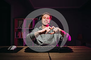 Happy guy gamer in a headset shows a heart gesture and looks at the camera with a smile on his face. Smiling streamer gamer