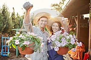 Happy guy and girl gardeners in a straw hats hold pots with wonderful petunia in the garden on a sunshine