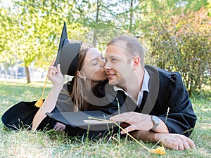 Happy guy and girl in graduate gowns lie on the lawn in the university park. A student girl in love kisses a guy on the