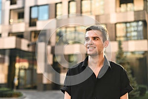 Happy guy model in stylish black shirt smiling on urban background, looking away at blank space. Handsome young man in shirt and