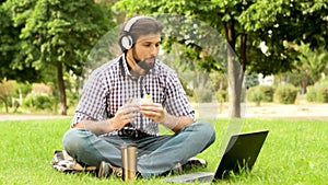 Happy guy is sitting on grass in oark and looking at laptop. He is listening to music and smiling. Guy is biting piece