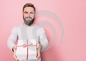 Happy guy is standing and holding a present. The box is white with red ribbon. Isolated on pink background.