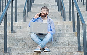 Happy guy talking on mobile phone working on laptop. Smiling guy making mobile call on stairs