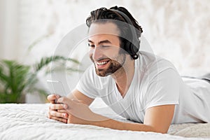 Happy guy using smartphone and headset, bedroom interior