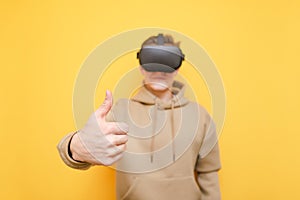 Happy guy in virtual reality helmet and casual clothes stands on a yellow background and shows thumbs up closeup. Young man would