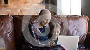 Happy hairless mother with little daughter using laptop, enjoying weekend