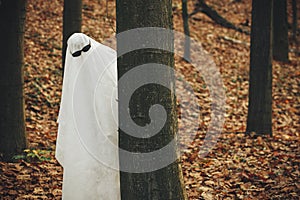 Happy Halloween! Funny ghost with black glasses peeking out of a tree in moody autumn forest. Person dressed with white sheet as