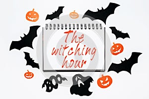 Happy halloween holiday concept. Notepad with text The witching hour on white background with bats, pumpkins and ghosts