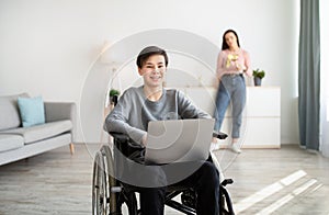 Happy handicapped teen boy in wheelchair with laptop computer indoors, his mother standing on background