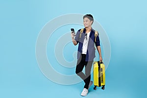 Happy handsome Asian tourist man holding smartphone with holding suitcase bag going to travel on holidays isolated on blue